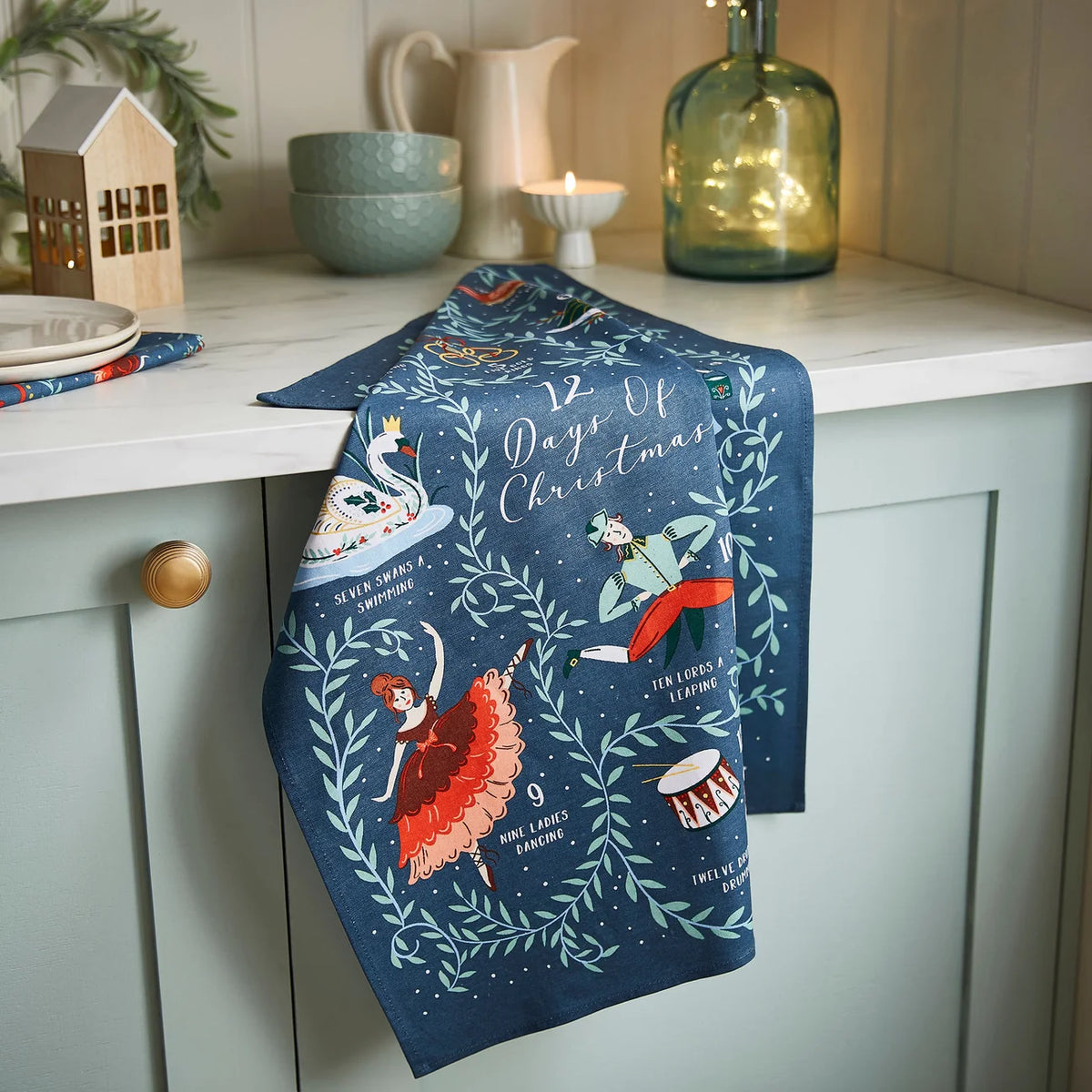 Festive cotton tea towel styled on a white marble kitchen counter next to Christmas decorations.