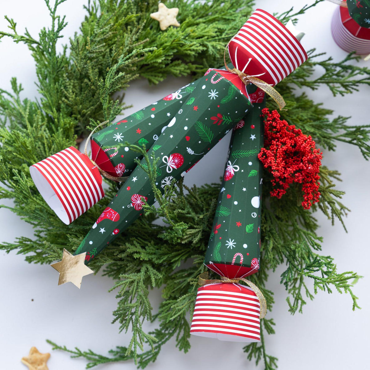 Three tree-shaped Christmas crackers with gold stars and striped bases arranged on a festive evergreen wreath.