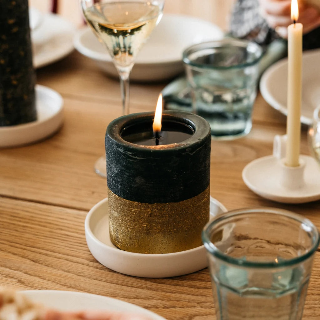 Lit gold-dipped Winter Thyme pillar candle on a white ceramic plate as part of a festive dining table setting.