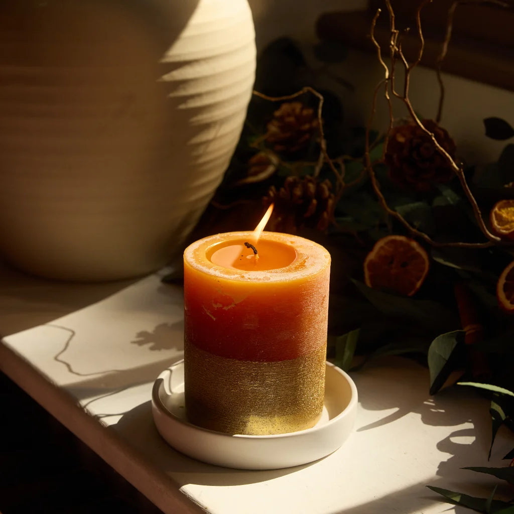 Lit gold-dipped orange candle on a white ceramic tray, styled with festive foliage and pine cones