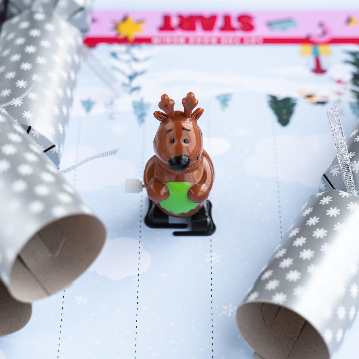 Reindeer wind-up toy positioned between two open silver Christmas crackers on a paper track