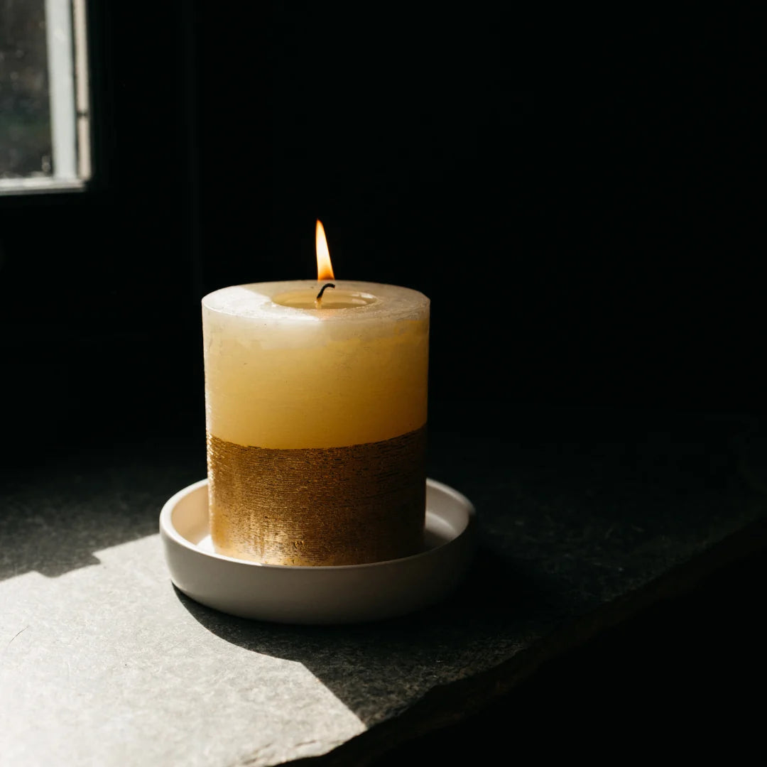 Lit Inspiritus gold-dipped pillar candle on a white ceramic tray by a window in low light.