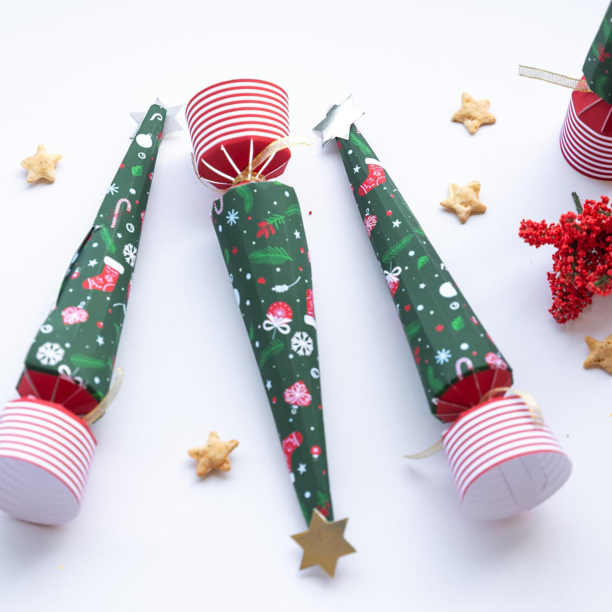 Three novelty tree crackers standing upright on a white surface with small star biscuits and red berries.