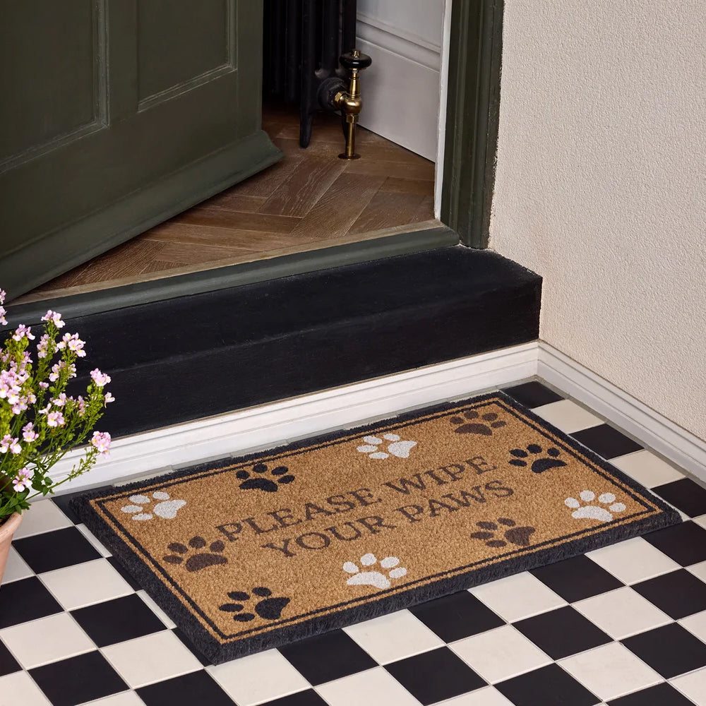 Wipe Your Paws doormat placed on a monochrome tiled floor in front of an open dark green doorway.