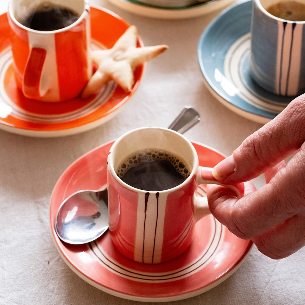 Lifestyle view of a rose stripe espresso mug being held over a matching saucer alongside orange and blue mugs.