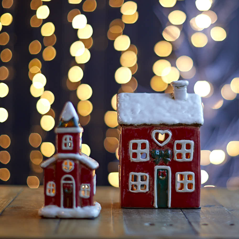 Festive ceramic house and church tea-light holders glowing on a wooden surface against blurred fairy lights.