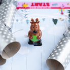 Reindeer wind-up toy positioned between two open silver Christmas crackers on a paper track