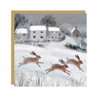 Dashing Hares Christmas card showing three hares running in snow towards cottages, with a gold border and red envelope.