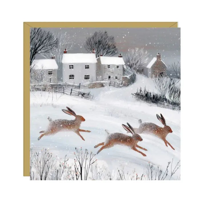 Dashing Hares Christmas card showing three hares running in snow towards cottages, with a gold border and red envelope.