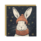 Hartley Christmas card illustration featuring a white rabbit wearing an orange knitted hat and scarf on a dark background.