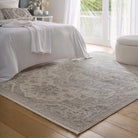 Lifestyle view of the neutral rug in a sunlit bedroom setting next to a white bed and footstool.