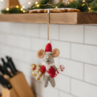 Festive felt mouse hanging from a wooden shelf in a kitchen setting with Christmas greenery in the background.