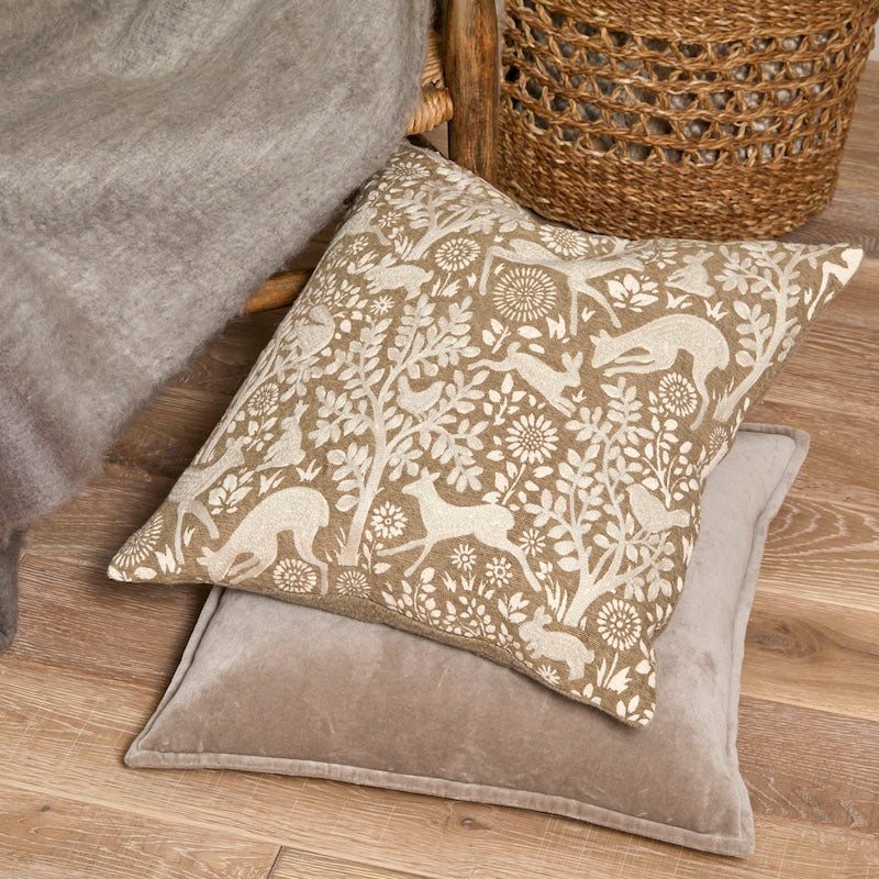 Embroidered woodland cushion styled on a wooden floor alongside a plain velvet cushion and wicker basket.