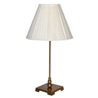 Slim table lamp with antique brass stem and square base, topped with a cream pleated shade against a white background.