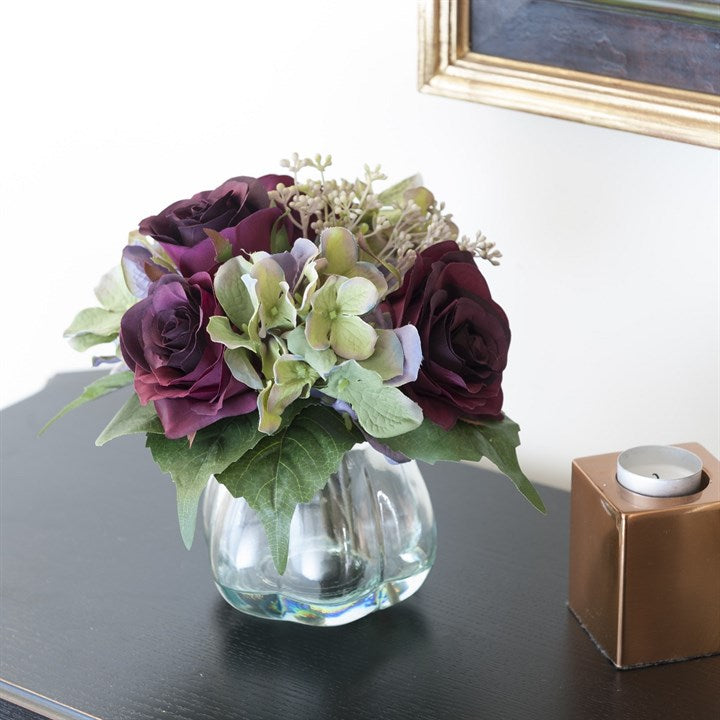 Faux floral arrangement of purple roses and hydrangeas in a glass vase styled on a dark wood table next to a candle