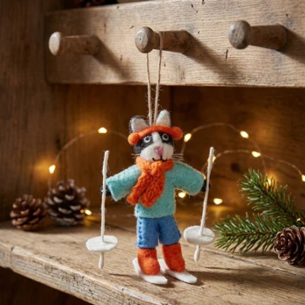 Felt skiing cat hanging from a wooden shelf decorated with pine cones and festive fairy lights