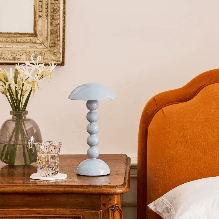 Lacquered blue bobbin lamp styled on a wooden bedside table beside a bed and glass vase
