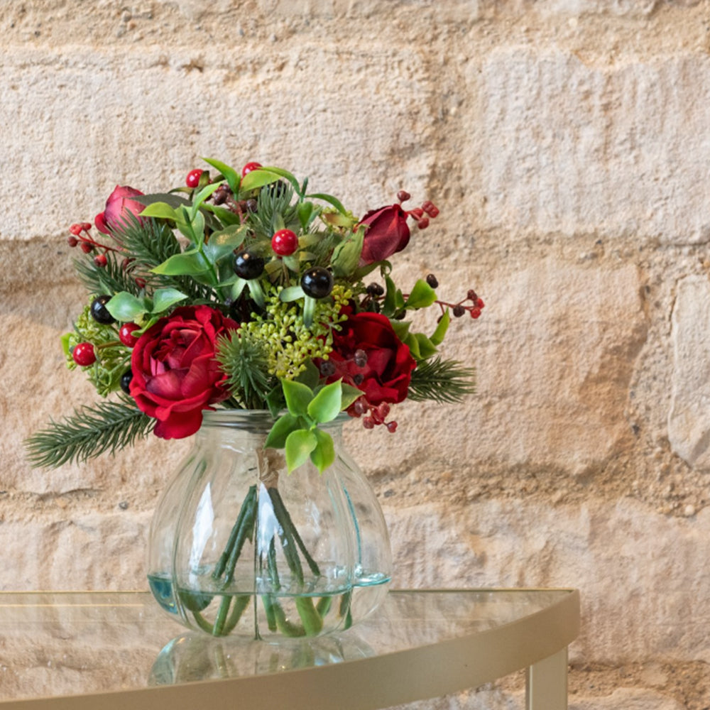 Faux rose and berry arrangement in a glass vase, styled on a gold side table against a stone wall.