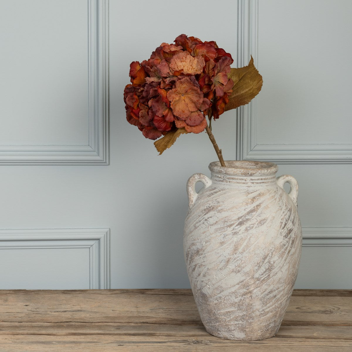 Single autumnal faux hydrangea stem displayed in a rustic cream ceramic vase on a wooden surface.
