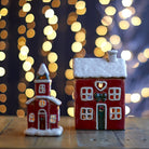 Festive ceramic house and church tea-light holders glowing on a wooden surface against blurred fairy lights.