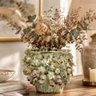 Ceramic floral pot styled on a wooden table with dried foliage and books in a warm interior setting.