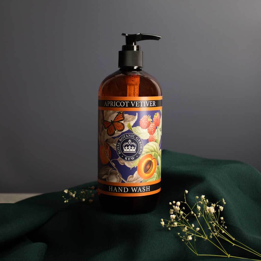 Kew Gardens Apricot Vetiver Hand Wash styled on a dark green fabric next to delicate white flowers.