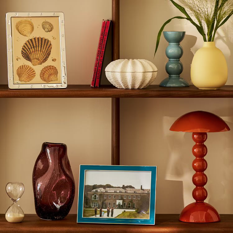 Small Chambray Blue candlestick styled on a wooden bookshelf alongside decorative bowls, frames, and an orange lamp.