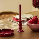 Small cherry red bobbin candlestick styled on a wooden dining table with a matching red candle and pomegranates.