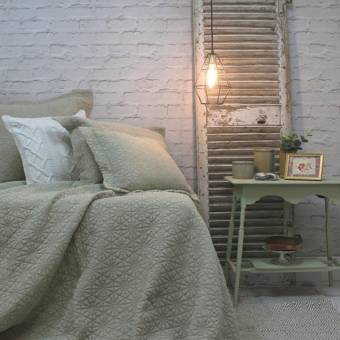 Pampas green quilted bedspread styled on a bed with matching pillows against a rustic white brick wall
